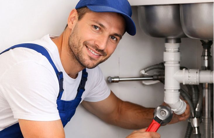 Klempner Göttingen und Umgebung Lächelnder Klempner in blauer Arbeitskleidung arbeitet an einem Rohr.