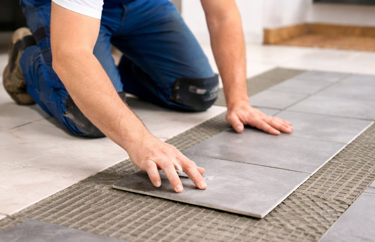 Reparatur- und Fliesenarbeiten in Göttingen und Umgebung Handwerker verlegt graue Fliesen auf einem Bodenbelag.