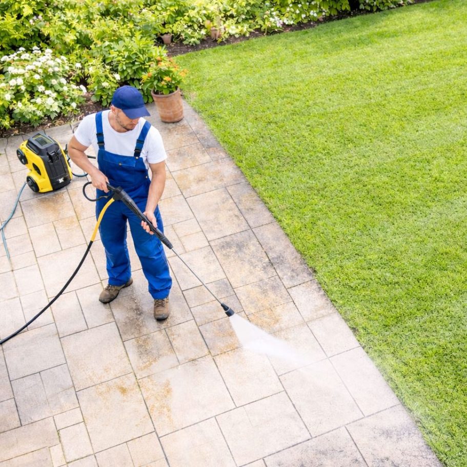 Garten- und Grundstückspflege Göttingen Mann in blauer Arbeitskleidung reinigt einen Steinboden mit einem Hochdruckreiniger.